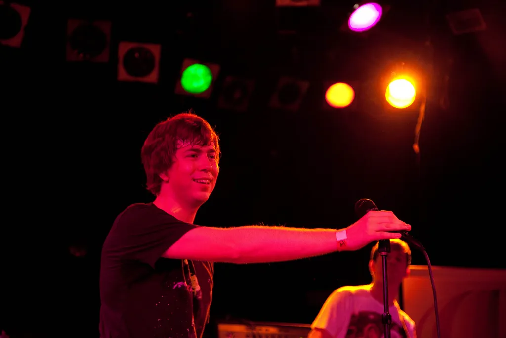 James Bourne smiling on stage, holding the microphone out towards the crowd under coloured lights