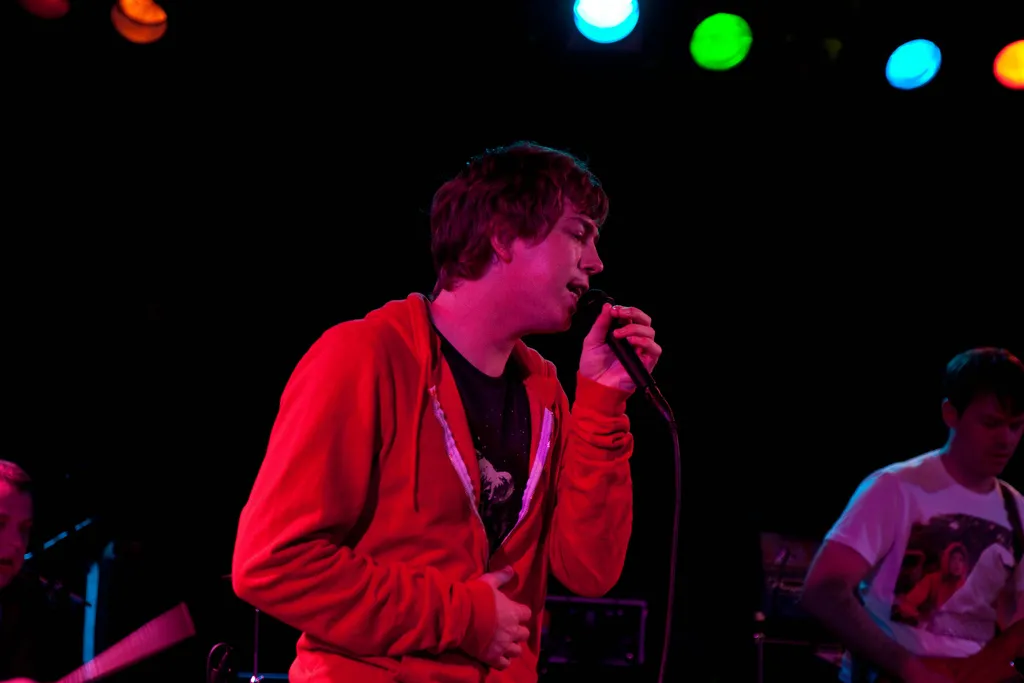 James Bourne singing into the microphone in a red hoodie, a guitarist to his right