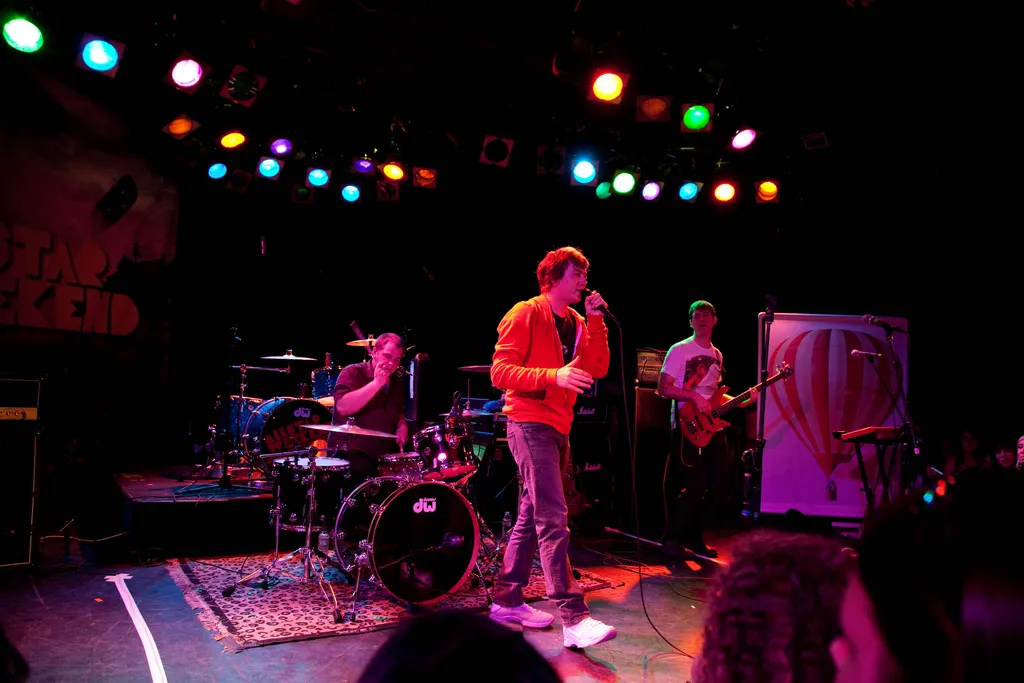 The band on stage under rows of coloured lights: James Bourne singing in a red hoodie, the drummer and guitarist behind him